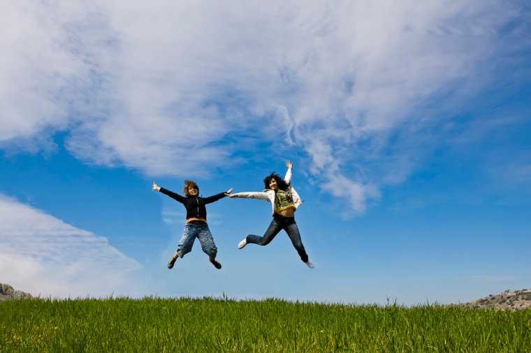 Feel Good Group - Two women in the sun jumping high above a field of green grass against a backdrop of blue sky. A physical change can create a change in energy